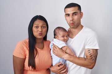 Fototapeta premium Young hispanic couple with baby standing together over isolated background puffing cheeks with funny face. mouth inflated with air, crazy expression.
