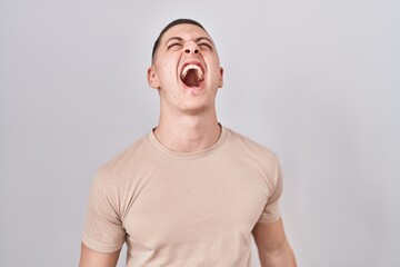 Young man standing over isolated background angry and mad screaming frustrated and furious,...