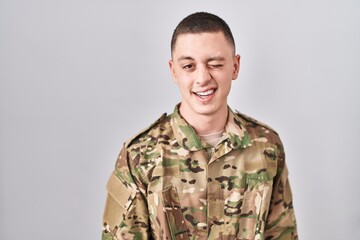 Young man wearing camouflage army uniform winking looking at the camera with sexy expression, cheerful and happy face.