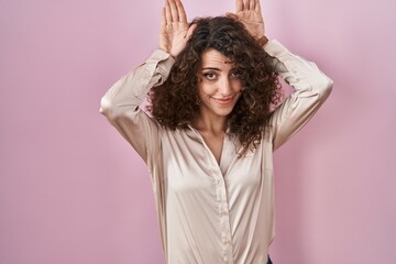 Naklejka premium Hispanic woman with curly hair standing over pink background doing bunny ears gesture with hands palms looking cynical and skeptical. easter rabbit concept.