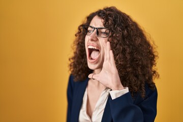 Hispanic woman with curly hair standing over yellow background shouting and screaming loud to side with hand on mouth. communication concept.