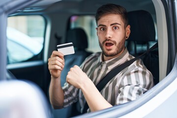 Hispanic man with beard driving car holding credit card scared and amazed with open mouth for surprise, disbelief face - obrazy, fototapety, plakaty