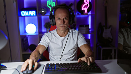 Fototapeta na wymiar Handsome middle age man streaming video game at night, sitting seriously concentrated at his cyber gaming room, fully equipped with futuristic technology