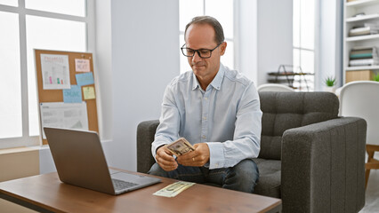 Confident middle age man, a happy business worker, basking in his success as he sits in office interior, counting dollars with a smile on his laptop