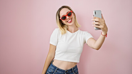 Young blonde woman wearing sunglasses make selfie by smartphone smiling over isolated pink...