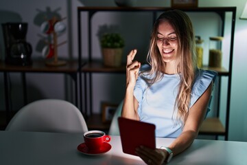 Young hispanic woman using touchpad sitting on the table at night gesturing finger crossed smiling with hope and eyes closed. luck and superstitious concept.