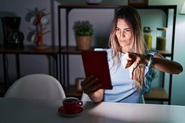 Young hispanic woman using touchpad sitting on the table at night looking unhappy and angry showing rejection and negative with thumbs down gesture. bad expression.