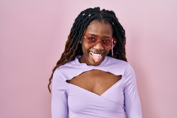 African woman with braided hair standing over pink background sticking tongue out happy with funny expression. emotion concept.