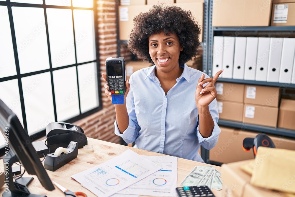 Poster Black woman with curly hair working at small business ecommerce holding credit card and dataphone smiling happy pointing with hand and finger to the side