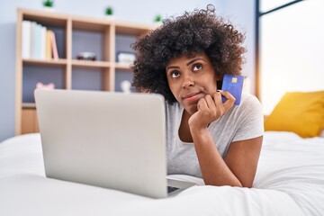 African american woman using laptop and credit card with doubt expression at bedroom
