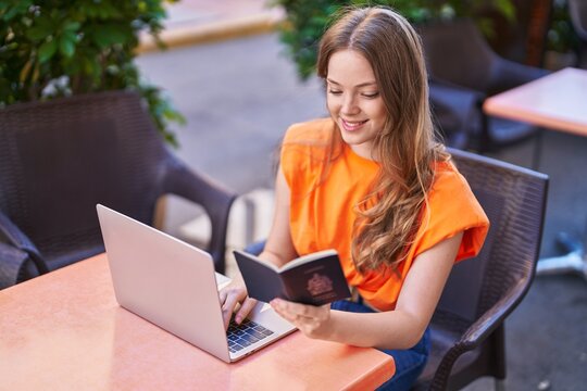 Young woman using laptop hiolding passport at coffee shop terrace