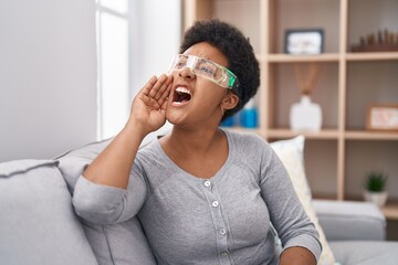 Young african american woman wearing virtual reality glasses sitting on the sofa clueless and...