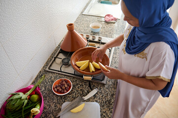 Authentic Muslim woman in hijab, standing by stove on kitchen counter and stacking potato slices in...