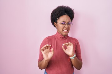 Beautiful african woman with curly hair standing over pink background disgusted expression,...