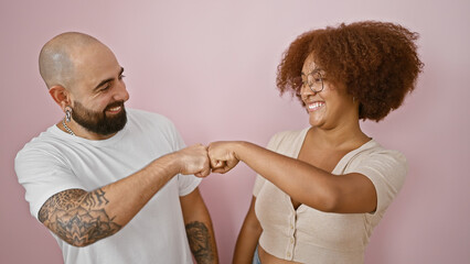 Beautiful couple standing together, bumping fists in celebration over an isolated pink background,...
