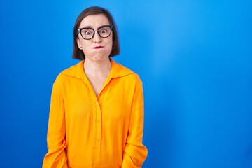 Middle age hispanic woman wearing glasses standing over blue background puffing cheeks with funny face. mouth inflated with air, crazy expression.