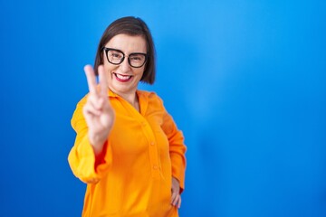 Middle age hispanic woman wearing glasses standing over blue background smiling looking to the camera showing fingers doing victory sign. number two.