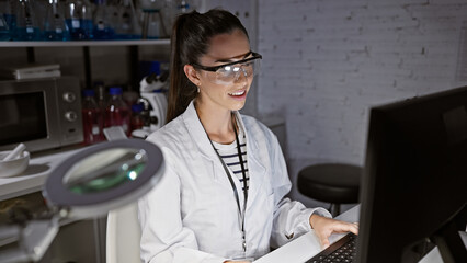 Smiling, confident young hispanic woman scientist, beautiful in her glasses, enjoying her work on the computer at the lab