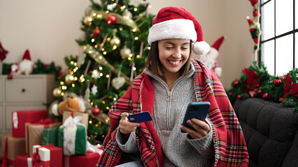 Young beautiful hispanic woman using smartphone and credit card sitting on sofa by christmas tree at home
