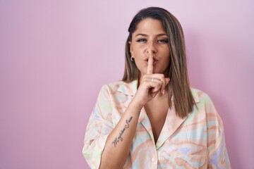 Blonde woman standing over pink background asking to be quiet with finger on lips. silence and...