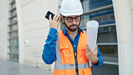Young hispanic man architect holding blueprints listening to voice message by smartphone at...
