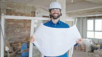 Young hispanic man architect reading house plans smiling at construction site