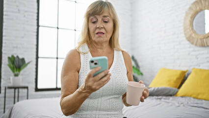 Fototapeta na wymiar Middle-aged blonde woman comfortably relaxed on bed, sipping coffee and texting on her phone, immersed in the serenity of her bedroom morning