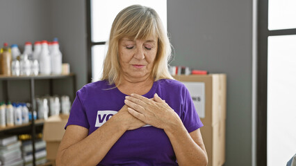 Fototapeta premium Middle age, blonde woman volunteer standing with love symbol gesture over chest in charity center, indoor, expressing lovely emotion.