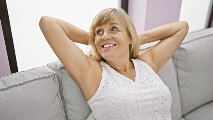 Joyful middle age blonde woman sitting comfortably on sofa at home, resting hands on head in a relaxed mood, exuding confidence and happiness.