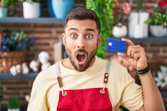 Handsome hispanic man working at florist shop holding credit card scared and amazed with open mouth for surprise, disbelief face