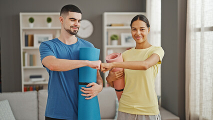 Obraz premium Beautiful couple bumping fists, holding a yoga mat, smiling with fitness glee in the home living room