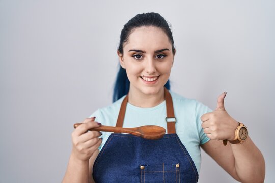 Young modern girl with blue hair wearing cook apron holding spoon smiling happy and positive, thumb up doing excellent and approval sign