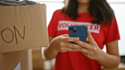 Tireless female volunteer's hands, a resilient hispanic woman, dutifully using her smartphone at heartwarming charity center amid boxes ready for donation packaging.