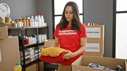 Selfless young hispanic woman volunteering at charity center, diligently packaging donated clothes with compassionate spirit, displaying beautiful altruism and unity