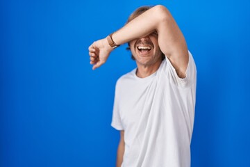 Middle age man standing over blue background covering eyes with arm smiling cheerful and funny. blind concept.