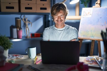 Middle age man sitting at art studio with laptop at night happy face smiling with crossed arms...