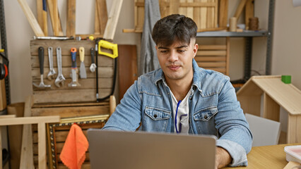 Serious-faced young hispanic man, a skilled carpenter, engrossed in his laptop amidst a busy carpentry workshop, presenting the blend of traditional woodwork and modern technology.