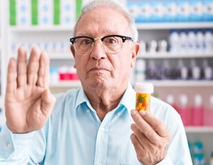 Senior man with grey hair holding pills at the pharmacy with open hand doing stop sign with serious and confident expression, defense gesture