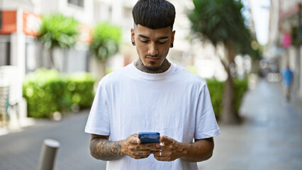 Cool and tattooed young latin man concentrated on his smartphone, texting while standing outdoors on a sunny city street sporting serious expression and relaxed lifestyle