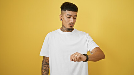 Handsome young latin man, tattooed and relaxed, looking at his wristwatch with a serious expression. he's standing against a cool, isolated yellow background, countdown timer in focus.