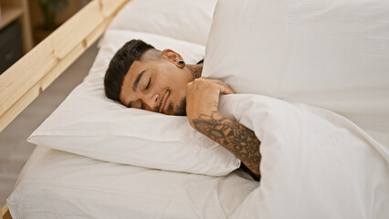 Handsome young latin man sleeping peacefully, lying in bed, covered by blankets in his apartment's comfortable bedroom, resting a tattooed body after a tiring morning