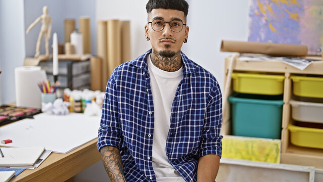 Serious-faced young latin man, tattooed artist in glasses, sitting indoors at art studio, engrossed in painting his canvas with passion