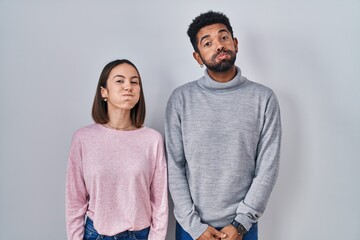 Young hispanic couple standing together puffing cheeks with funny face. mouth inflated with air, crazy expression.