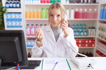 Young caucasian woman working at pharmacy drugstore speaking on the telephone doing money gesture with hands, asking for salary payment, millionaire business