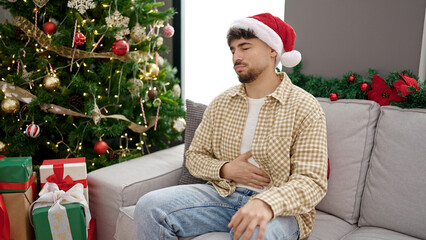 Young arab man suffering stomach ache sitting by christmas tree at home