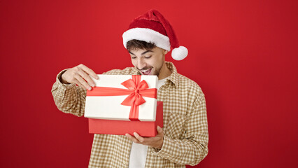 Young arab man wearing christmas hat unpacking gift over isolated red background