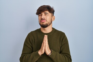 Arab man with beard standing over blue background begging and praying with hands together with hope...