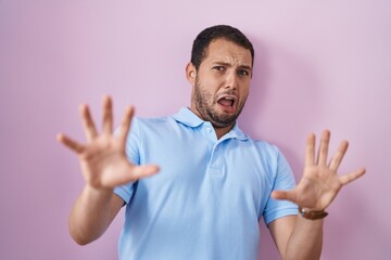 Hispanic man standing over pink background afraid and terrified with fear expression stop gesture with hands, shouting in shock. panic concept.