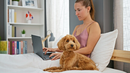 Young caucasian woman with dog using laptop sitting on bed at bedroom