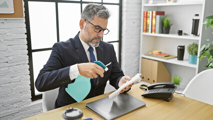 Handsome, grey-haired hispanic man cleaning his laptop at the office as part of his daily...